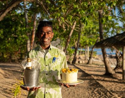 Smiling man in a tropical shirt holds a bucket with champagne and a decorated cake on a beach, surrounded by trees and dappled sunlight, evoking a celebratory and joyful atmosphere.