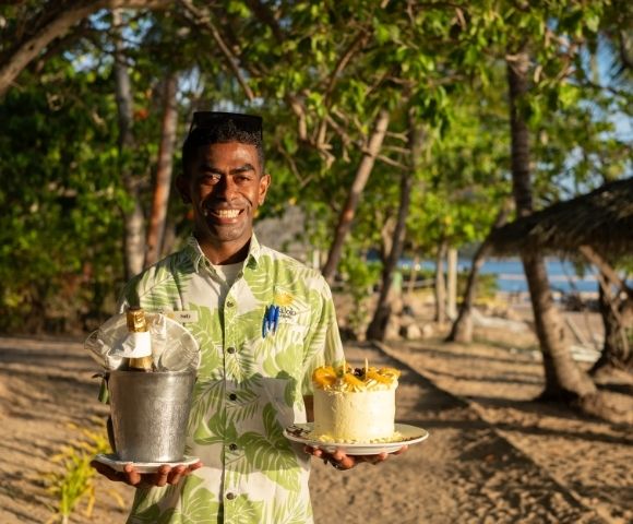 Smiling man in a tropical shirt holds a bucket with champagne and a decorated cake on a beach, surrounded by trees and dappled sunlight, evoking a celebratory and joyful atmosphere.