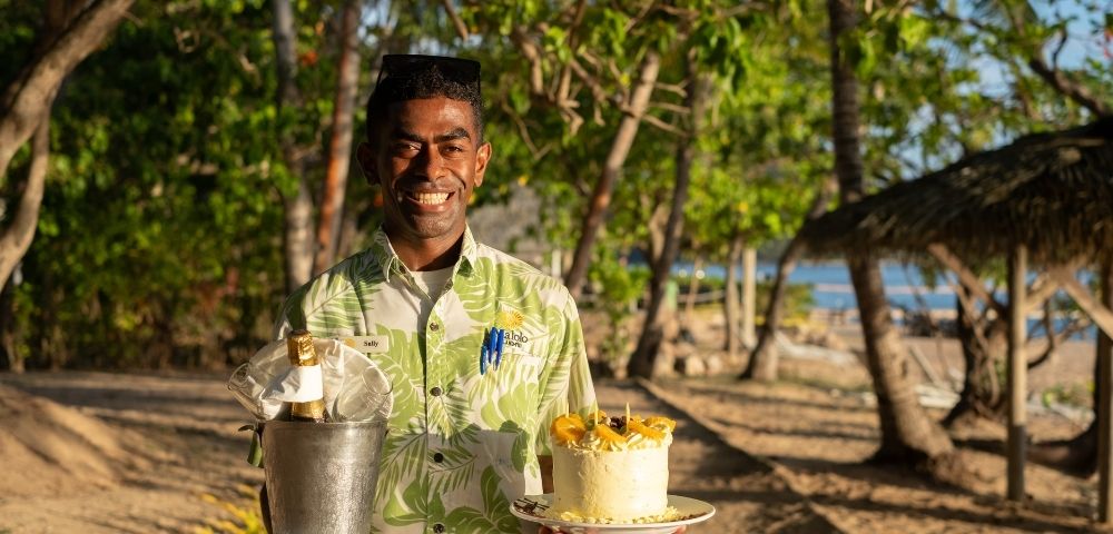 Smiling man in a tropical shirt holds a bucket with champagne and a decorated cake on a beach, surrounded by trees and dappled sunlight, evoking a celebratory and joyful atmosphere.