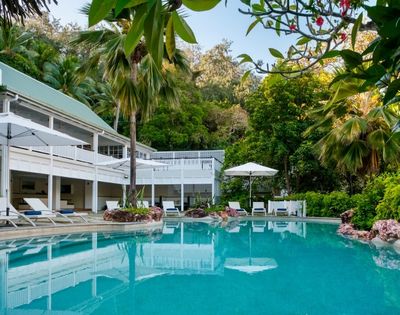 Luxurious tropical pool with clear turquoise water surrounded by lush greenery. A white modern house with balconies and poolside loungers under umbrellas adds elegance.