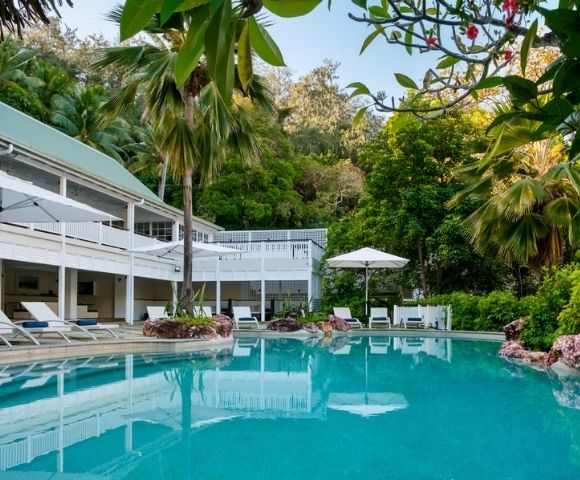 Luxurious tropical pool with clear turquoise water surrounded by lush greenery. A white modern house with balconies and poolside loungers under umbrellas adds elegance.