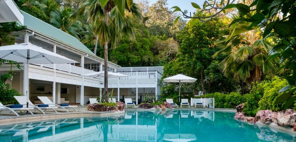 Luxurious tropical pool with clear turquoise water surrounded by lush greenery. A white modern house with balconies and poolside loungers under umbrellas adds elegance.
