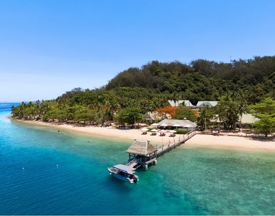 Aerial view of a serene beach resort with turquoise water, white sand, lush palm trees, and a pier with a boat docked. Clear blue sky above.