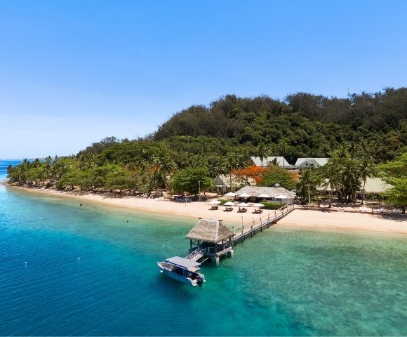 Aerial view of a serene beach resort with turquoise water, white sand, lush palm trees, and a pier with a boat docked. Clear blue sky above.