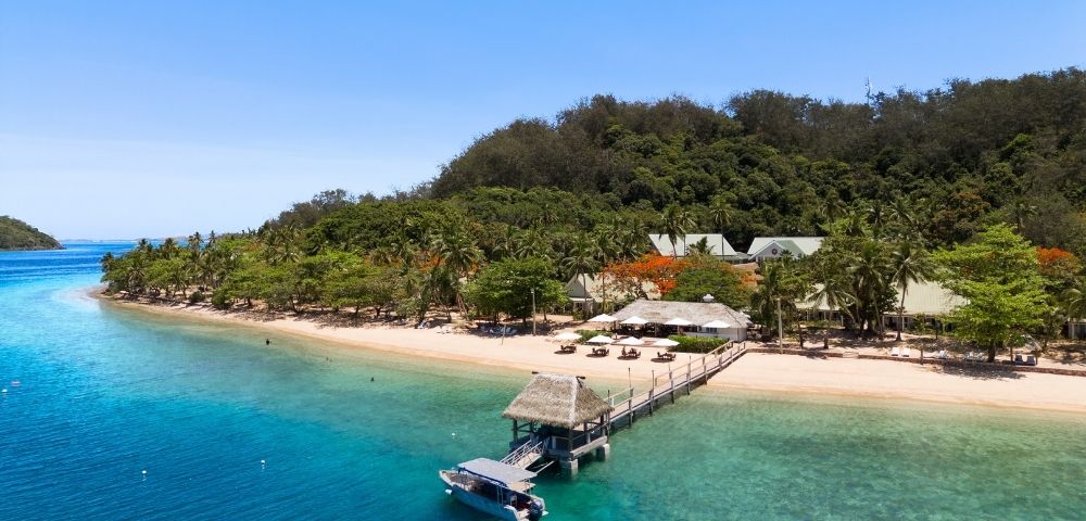 Aerial view of a serene beach resort with turquoise water, white sand, lush palm trees, and a pier with a boat docked. Clear blue sky above.
