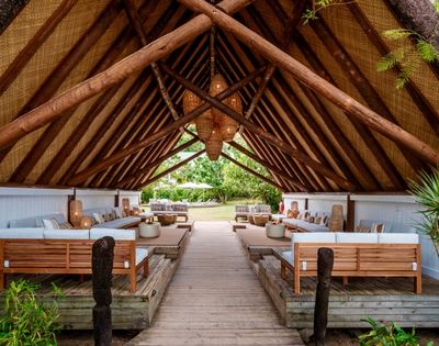 Rustic open-air pavilion with a thatched roof, featuring wooden lounge furniture and a central path lined with lush greenery. Relaxing, tropical ambiance.