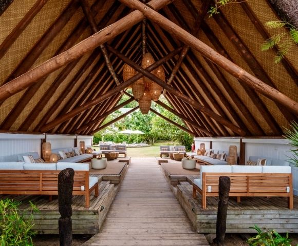 Rustic open-air pavilion with a thatched roof, featuring wooden lounge furniture and a central path lined with lush greenery. Relaxing, tropical ambiance.