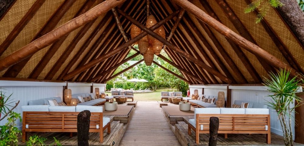 Rustic open-air pavilion with a thatched roof, featuring wooden lounge furniture and a central path lined with lush greenery. Relaxing, tropical ambiance.
