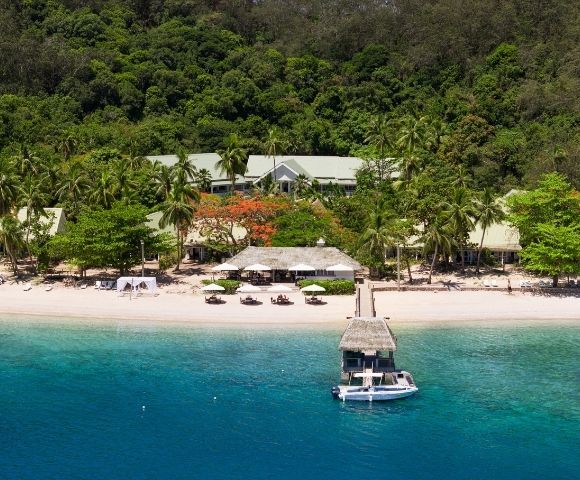 Aerial view of a serene tropical beach with lush palm trees and vibrant greenery. Sandy shore with sun loungers and a docked boat in clear blue water.