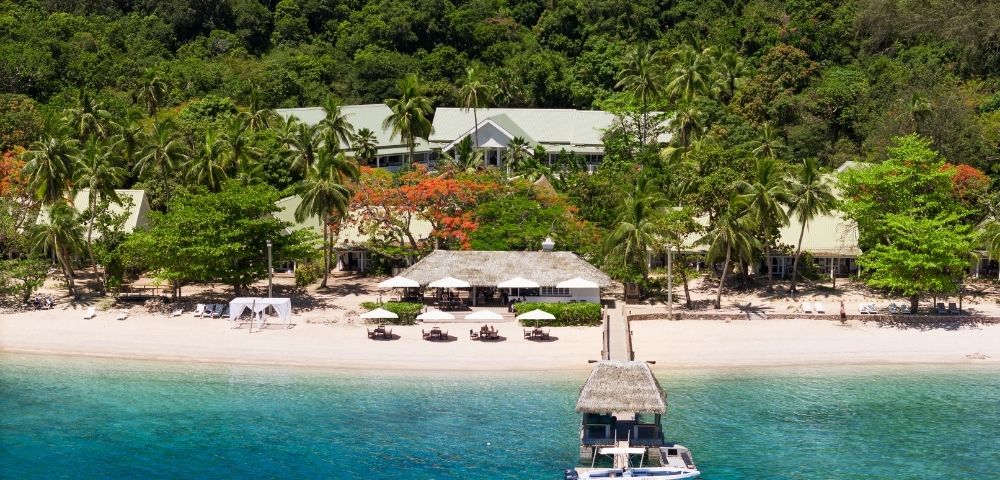Aerial view of a serene tropical beach with lush palm trees and vibrant greenery. Sandy shore with sun loungers and a docked boat in clear blue water.