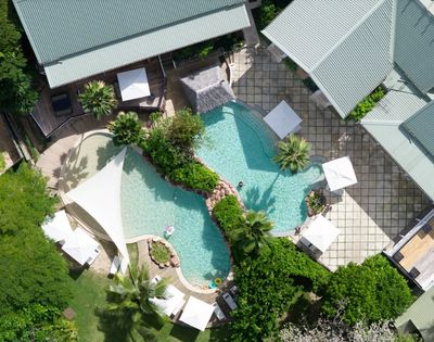 Aerial view of a luxury backyard with a uniquely-shaped pool surrounded by lush greenery, lounge chairs, white umbrellas, and modern buildings. Relaxing vibe.