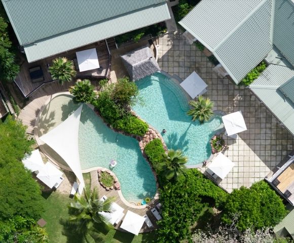 Aerial view of a luxury backyard with a uniquely-shaped pool surrounded by lush greenery, lounge chairs, white umbrellas, and modern buildings. Relaxing vibe.