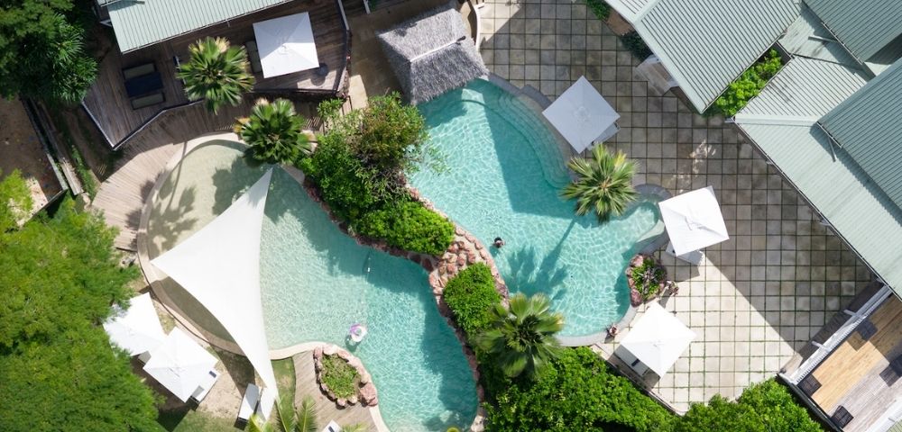 Aerial view of a luxury backyard with a uniquely-shaped pool surrounded by lush greenery, lounge chairs, white umbrellas, and modern buildings. Relaxing vibe.