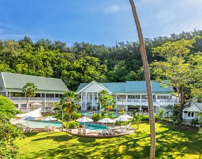 Serene resort with green-roofed buildings, lush trees, and a sunlit pool surrounded by umbrellas and loungers. Calm, inviting atmosphere.
