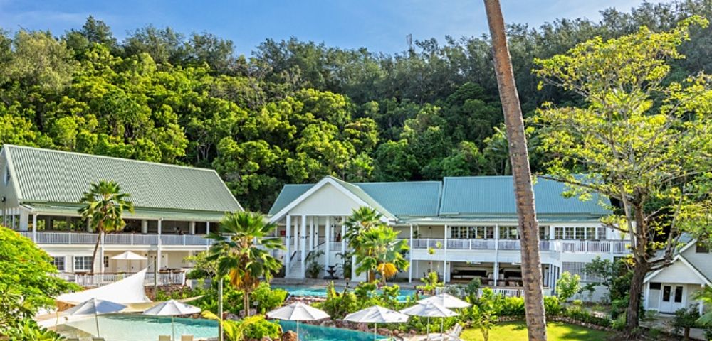 Serene resort with green-roofed buildings, lush trees, and a sunlit pool surrounded by umbrellas and loungers. Calm, inviting atmosphere.