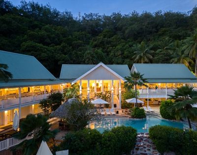 Aerial view of an illuminated tropical resort at dusk, featuring a central pool surrounded by lush greenery and buildings with green roofs, creating a serene ambiance.