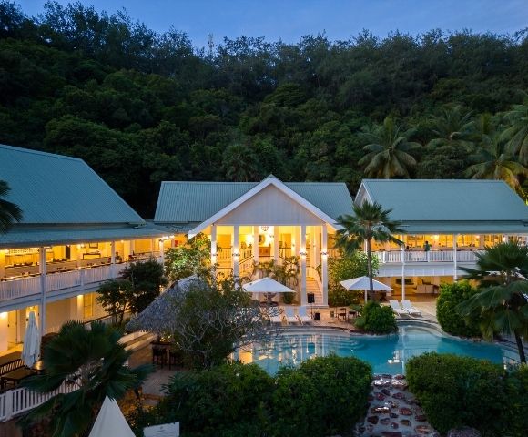 Aerial view of an illuminated tropical resort at dusk, featuring a central pool surrounded by lush greenery and buildings with green roofs, creating a serene ambiance.