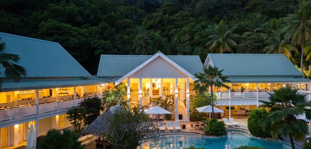 Aerial view of an illuminated tropical resort at dusk, featuring a central pool surrounded by lush greenery and buildings with green roofs, creating a serene ambiance.