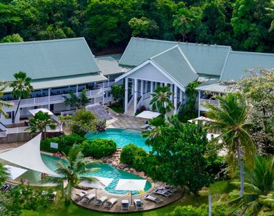 Aerial view of a tropical resort with a central pool, surrounded by lush greenery and palm trees. White buildings have green roofs, and lounge chairs are poolside.