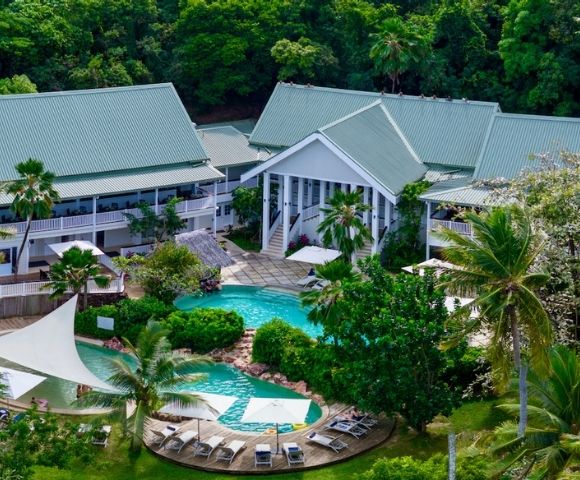 Aerial view of a tropical resort with a central pool, surrounded by lush greenery and palm trees. White buildings have green roofs, and lounge chairs are poolside.