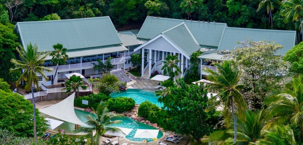 Aerial view of a tropical resort with a central pool, surrounded by lush greenery and palm trees. White buildings have green roofs, and lounge chairs are poolside.