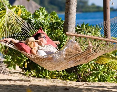 A couple relaxes in a hammock on a tropical beach, surrounded by lush greenery and sunlight. The serene ocean and palm trees create a peaceful, idyllic scene.