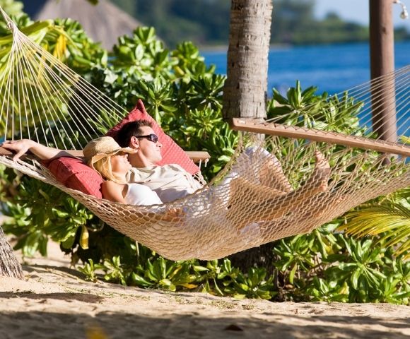 A couple relaxes in a hammock on a tropical beach, surrounded by lush greenery and sunlight. The serene ocean and palm trees create a peaceful, idyllic scene.