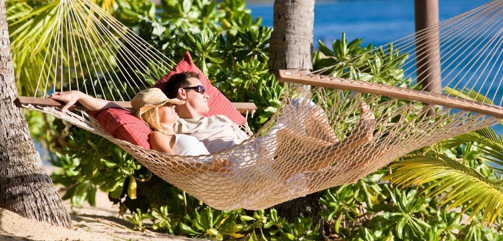 A couple relaxes in a hammock on a tropical beach, surrounded by lush greenery and sunlight. The serene ocean and palm trees create a peaceful, idyllic scene.