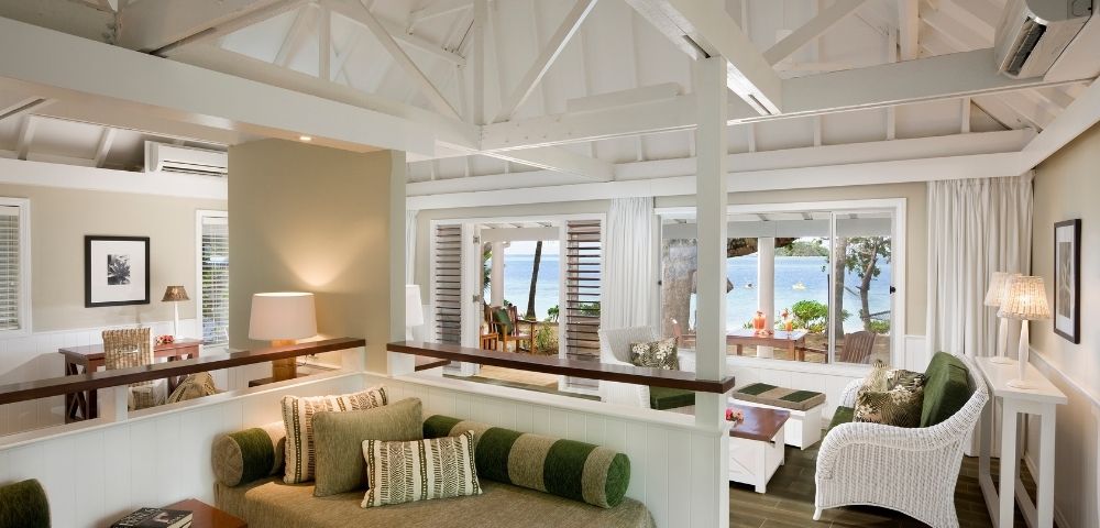 Airy coastal living room with white wicker furniture and green accents. Large windows offer a serene ocean view, conveying relaxation and tranquility.