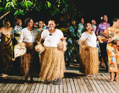 A group of people in traditional attire, including grass skirts and white tops, dance joyfully on a wooden deck at night, surrounded by greenery and a festive atmosphere.