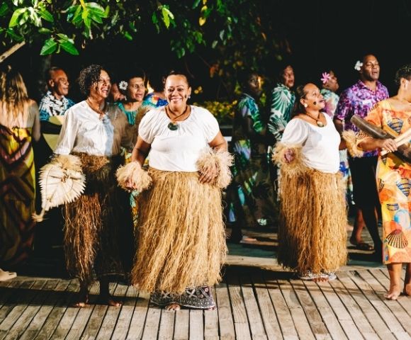 A group of people in traditional attire, including grass skirts and white tops, dance joyfully on a wooden deck at night, surrounded by greenery and a festive atmosphere.