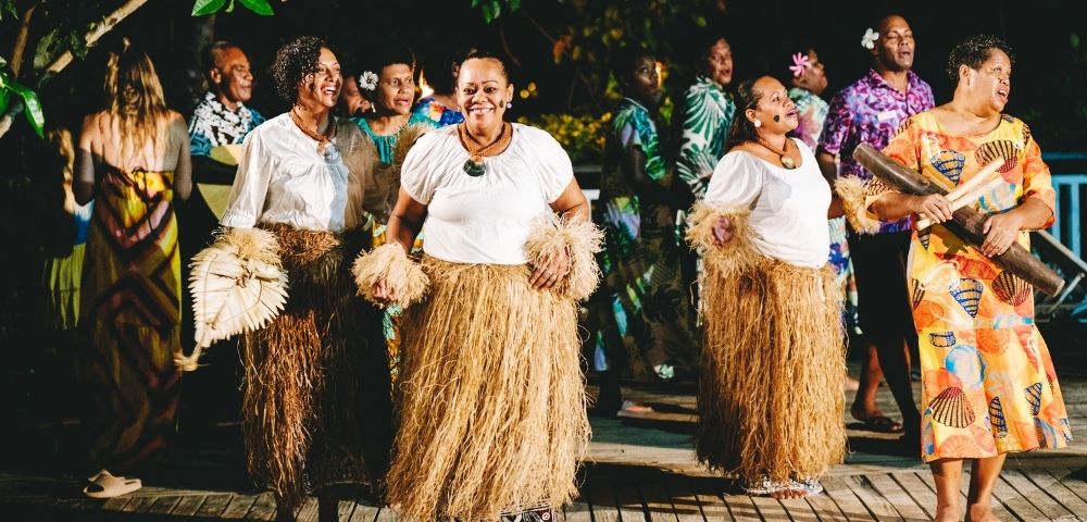 A group of people in traditional attire, including grass skirts and white tops, dance joyfully on a wooden deck at night, surrounded by greenery and a festive atmosphere.