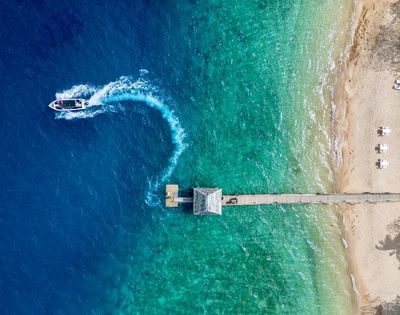 Aerial view of a pier extending into gradient blue to green sea. A boat creates a white wake in the water. Sandy beach with loungers on the right.