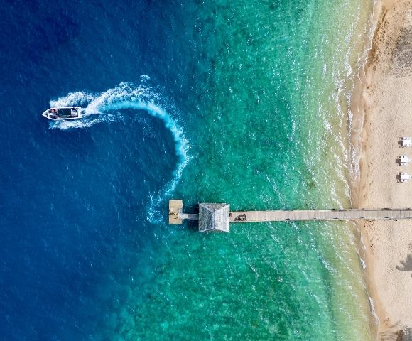 Aerial view of a pier extending into gradient blue to green sea. A boat creates a white wake in the water. Sandy beach with loungers on the right.