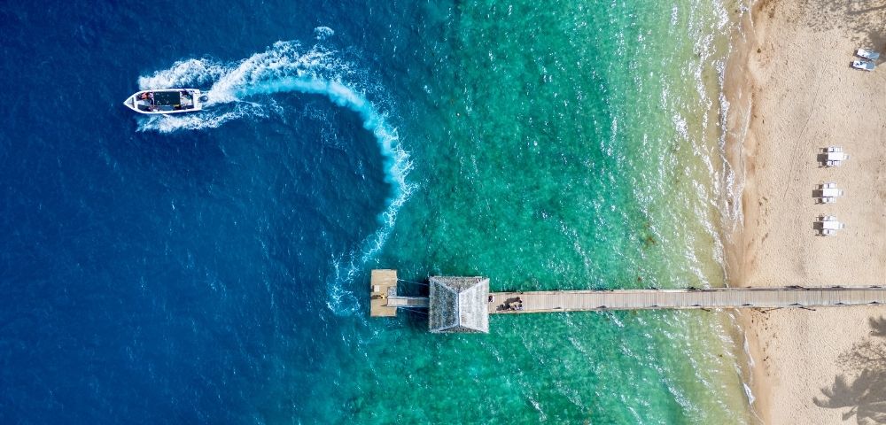 Aerial view of a pier extending into gradient blue to green sea. A boat creates a white wake in the water. Sandy beach with loungers on the right.