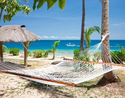 A hammock tied between palm trees on a sandy beach overlooks a calm turquoise sea. A thatched umbrella and a small boat enhance the serene tropical setting.