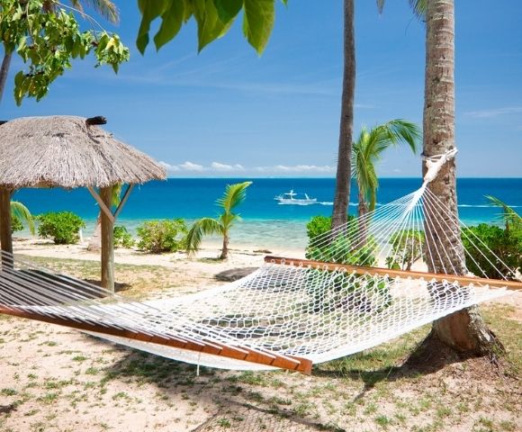 A hammock tied between palm trees on a sandy beach overlooks a calm turquoise sea. A thatched umbrella and a small boat enhance the serene tropical setting.