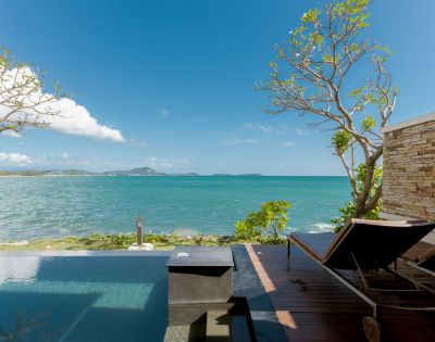 A serene beachfront view with an infinity pool, wooden deck, and two loungers. Clear blue sky and distant islands evoke a tranquil, relaxing atmosphere.