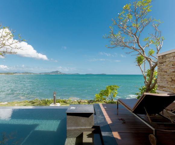A serene beachfront view with an infinity pool, wooden deck, and two loungers. Clear blue sky and distant islands evoke a tranquil, relaxing atmosphere.