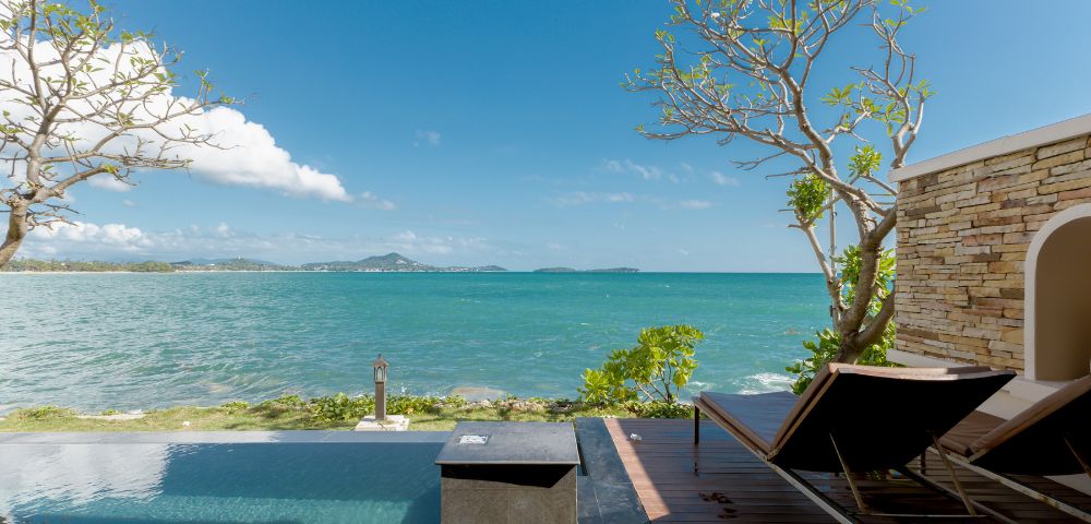 A serene beachfront view with an infinity pool, wooden deck, and two loungers. Clear blue sky and distant islands evoke a tranquil, relaxing atmosphere.