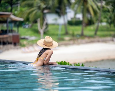 Woman in a straw hat leans on the edge of an infinity pool, gazing at a serene beach with palm trees, conveying relaxation and tropical vibes.