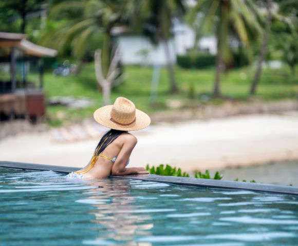 Woman in a straw hat leans on the edge of an infinity pool, gazing at a serene beach with palm trees, conveying relaxation and tropical vibes.