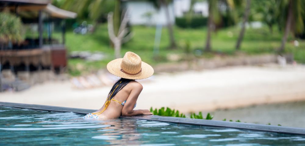 Woman in a straw hat leans on the edge of an infinity pool, gazing at a serene beach with palm trees, conveying relaxation and tropical vibes.