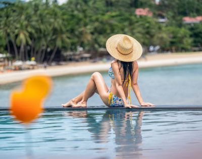 A woman in a straw hat and floral bikini sits by an infinity pool, facing the ocean. Palm trees and blurred beach background convey a tropical, relaxing vibe.