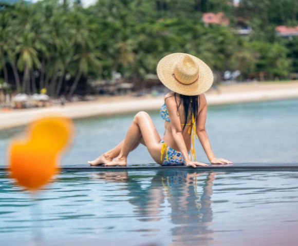 A woman in a straw hat and floral bikini sits by an infinity pool, facing the ocean. Palm trees and blurred beach background convey a tropical, relaxing vibe.