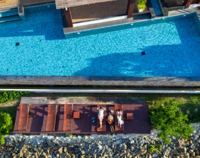 Aerial view of two people relaxing on lounge chairs by a clear blue pool, surrounded by greenery and stones, under a sunny, serene sky.