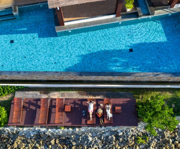 Aerial view of two people relaxing on lounge chairs by a clear blue pool, surrounded by greenery and stones, under a sunny, serene sky.