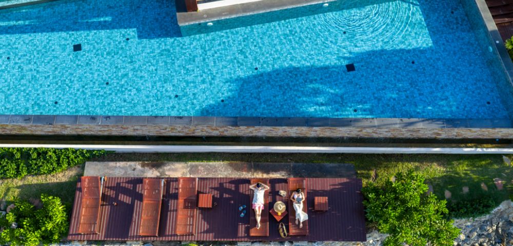Aerial view of two people relaxing on lounge chairs by a clear blue pool, surrounded by greenery and stones, under a sunny, serene sky.