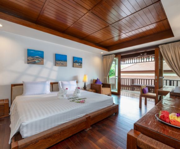 Elegant hotel room featuring a large bed with white linens, wooden furniture, artwork on the wall, and a balcony with open curtains letting in natural light.