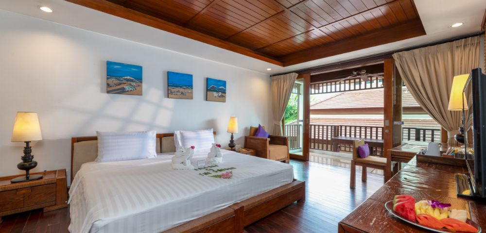 Elegant hotel room featuring a large bed with white linens, wooden furniture, artwork on the wall, and a balcony with open curtains letting in natural light.